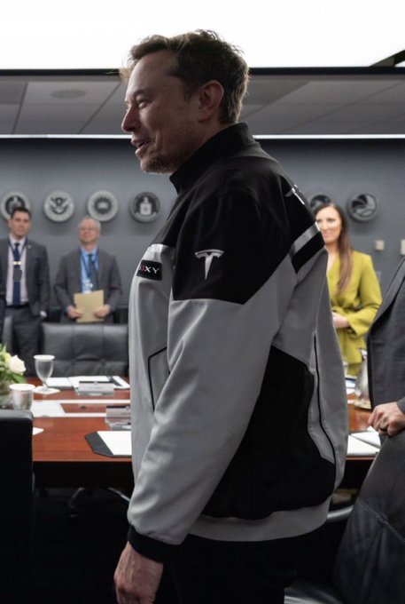 I'm not able to identify the people in this image based on their facial features. However, I can describe what I see:

A man wearing a black and gray Tesla jacket with the company logo on the chest stands in what appears to be a corporate meeting room, with other people visible in the background, decorative seals or emblems mounted on the wall, and papers spread across a conference table.