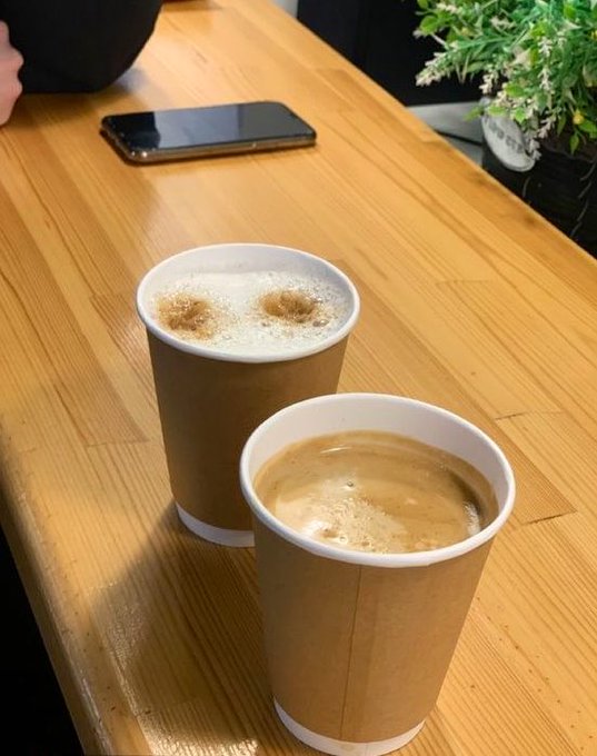 Two paper coffee cups sit on a wooden table, one with foam art and another with latte, alongside a smartphone in the background.