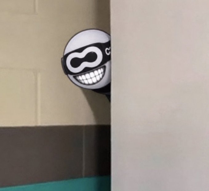 A cartoonish "vibe check" meme character with a masked smile and the text "vi" peeks around a corner in what appears to be a restroom setting. The image's playful peeking pose humorously illustrates the post's theme of "looking deeper" into the technical differences between Optimum's mump2p and Flexnodes, as if the meme is sneaking a curious glance to discover the hidden details.