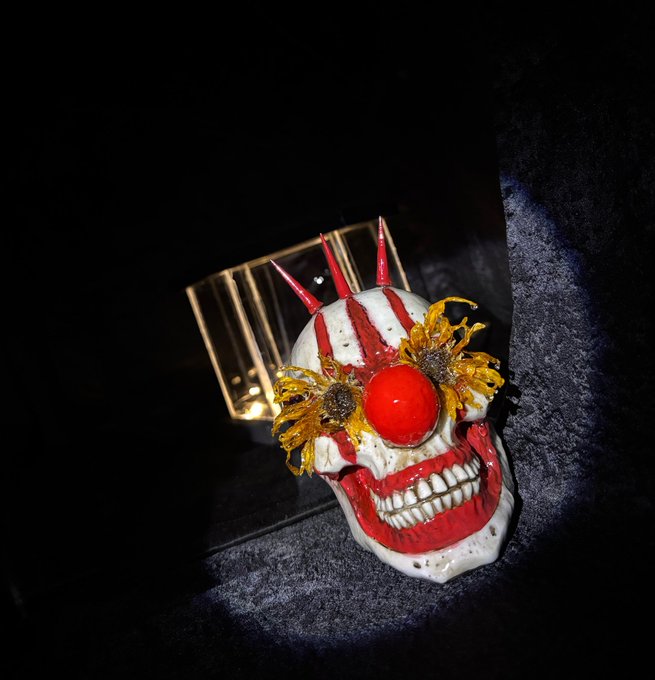 A close-up photograph of a decorative ceramic skull painted white with red accents, featuring a red spherical nose, yellow sunflower decorations over the eyes, and red horn-like protrusions on top, resting on a dark surface.