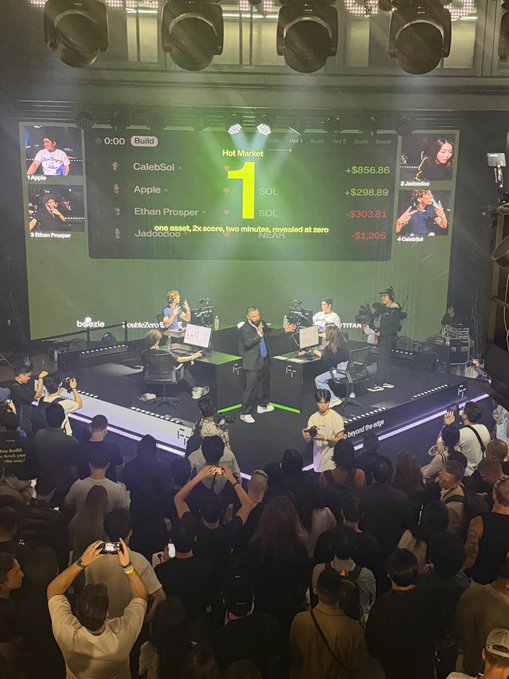A live event stage features large screens displaying a 'Hot Market' leaderboard showing crypto tickers like 'SOL' with prices and player handles such as 'CalebSol' and 'Apple', while people on stage interact with equipment before an audience.
