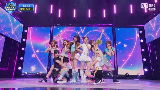 A vibrant stage performance showing a group of nine female performers posing together under pink lighting, with 'M COUNTDOWN' and 'Mnet' logos visible on screen.