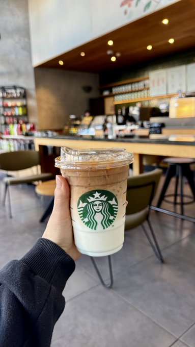 A hand holds up a plastic Starbucks cup containing a layered iced coffee drink, photographed inside a cafe with a counter and seating visible in the background.