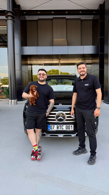Two men pose proudly in front of a black Mercedes-Benz with the personalized license plate "07 BTC 001," celebrating the new plate with a small dog held by the man on the left. The custom plate cleverly references Bitcoin's ticker symbol (BTC), aligning with the congratulatory post about this unique crypto-themed vanity plate.