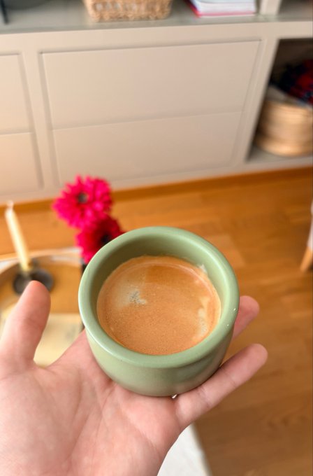 A hand holds a small green ceramic cup containing an espresso, with white cabinetry and pink flowers visible in the background.