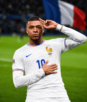 Kylian Mbappé, wearing the white France #10 jersey, salutes with his hand to his temple on the pitch with a French flag visible in the background, visually representing the French side of the high-stakes France vs. Morocco match that a Polymarket trader bet $5.14 million on to exceed 2.5 goals.