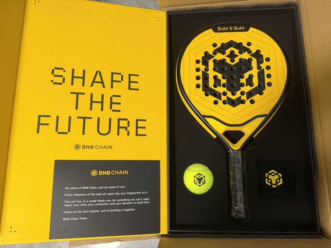 A BNB Chain 'Shape the Future' gift box containing a yellow padel racket labeled 'Build N Build' along with a tennis ball and a branded card celebrating six years of BNB Chain.