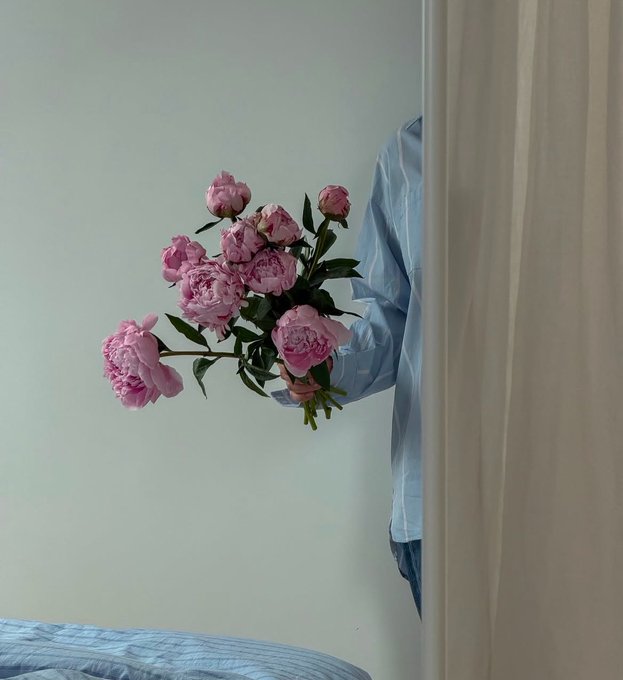 The image features a person holding a bouquet of pink flowers against a white wall. The background shows a bed covered with blue sheets. The photo has been captioned, "Happy Sunday My babies" and "Remember @TheARCTERMINAL Has Most forecasting tools & they give you a single number".