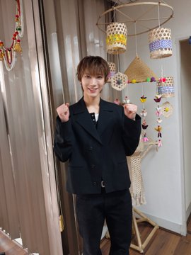 A young man in a black blazer smiles and makes double fists in a celebratory pose inside a room decorated with a colorful paper-craft mobile and a straw hat. The hashtags "#TEN" and "#WTCE2026" suggest the image captures a joyful moment related to the TEN group, possibly marking excitement around the 2026 World Tour.