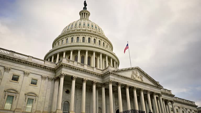 The image shows a large white building with columns and a dome, which appears to be the United States Capitol Building. A flag can be seen flying from the top of the building. In addition to these features, there are several other buildings visible in the background that contribute to the overall scene.