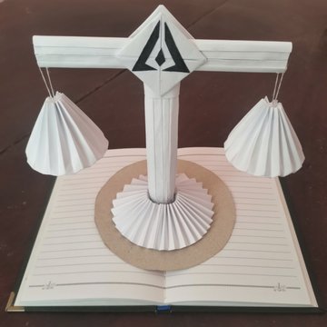 A pop-up paper craft sculpture depicting a balance scale with a logo at the top, rising from the center of an open book.