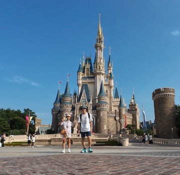 A photograph of two people standing in front of Cinderella's Castle at a Disney theme park on a sunny day with a clear blue sky.