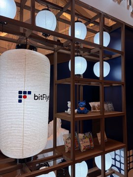 A row of white paper lanterns featuring the "bitFlyer" logo hangs in a wooden display alongside plush toys and magazines, reflecting the cryptocurrency exchange's presence at the WebX 2026 conference.
