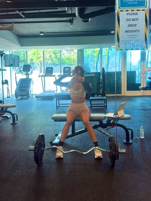 A woman in a beige two-piece workout set takes a mirror selfie in a gym while seated on a bench with an EZ curl bar loaded with weights.
