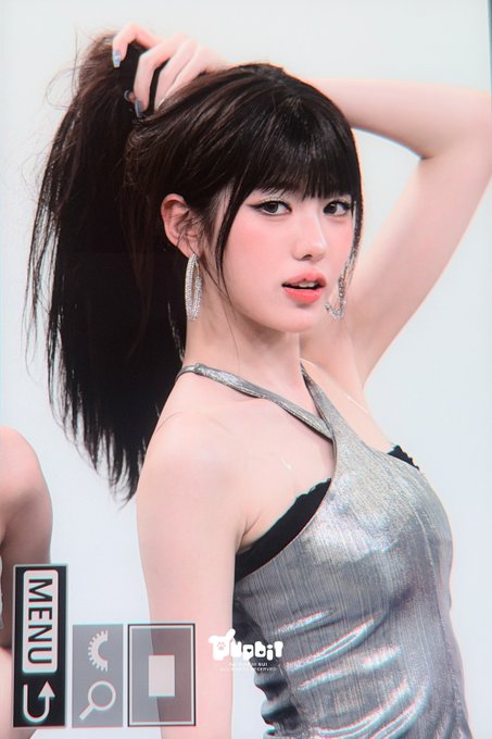 A young woman with dark hair in a high ponytail wearing a silver metallic halter top and dangling earrings, posing with her arm raised.