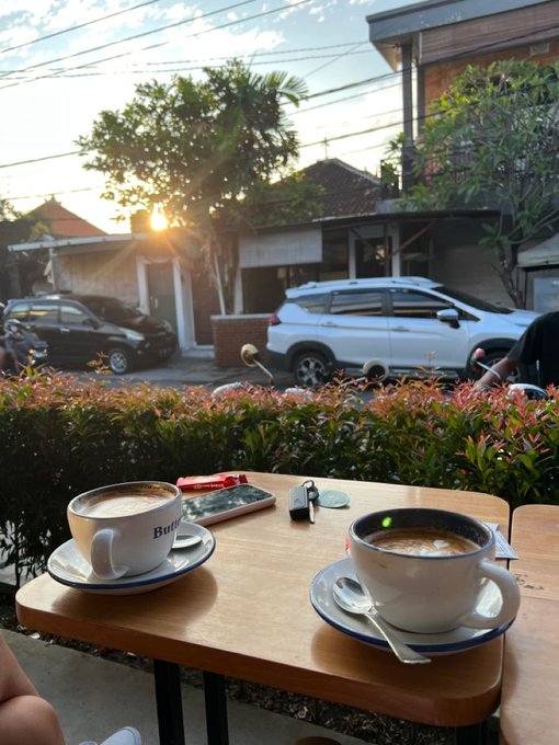 A first-person view of two coffee cups on a wooden cafe table at sunset, with parked cars and traditional buildings visible along the street in the background.
