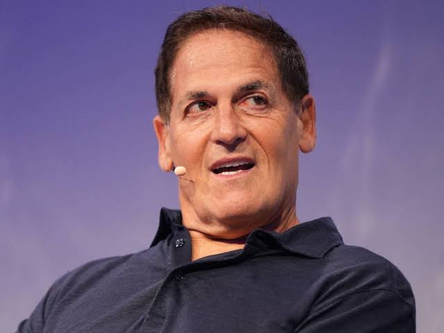 A man with short dark hair wearing a navy polo shirt and a headset microphone speaks at what appears to be a conference or presentation event.