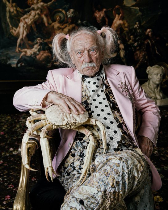 An elderly man with pigtails wears a pink blazer and patterned outfit while seated, holding a large crab on a golden chair with classical paintings in the background.