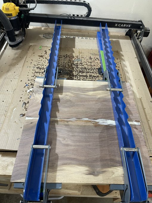 An X-Carve CNC router machine on a workbench holding a piece of wood secured with blue clamps and a smaller wooden board.
