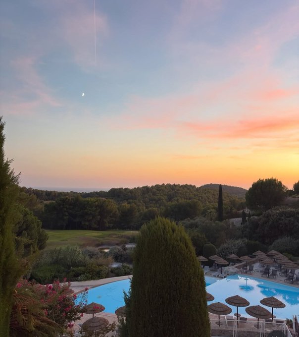 Scenic sunset view over a resort pool area surrounded by trees and Mediterranean landscape, with a crescent moon visible in the colorful sky.