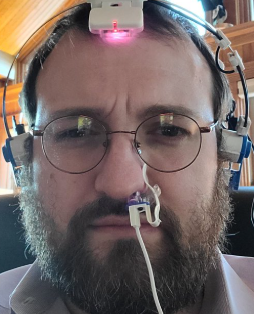 The image features a man with glasses, a beard, and a goatee wearing headphones that are connected to his face. He has a serious expression as he looks directly at the camera. The caption below the photo reads "Crapdano community right now:". This suggests that the man is participating in an online event or gathering related to Crapdano, possibly discussing or sharing information about the topic of interest with others through video conferencing platforms.