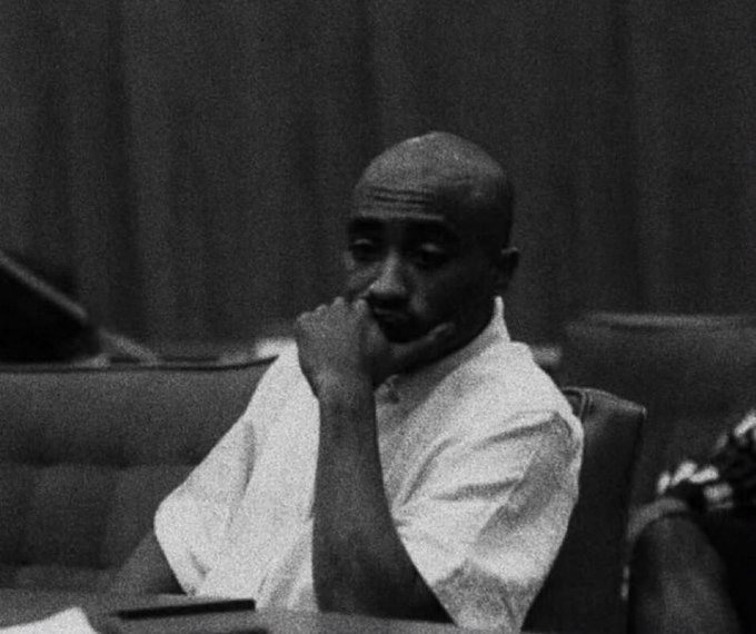 A black-and-white photo of a young man in a white shirt sitting in a chair, resting his chin thoughtfully on his hand.