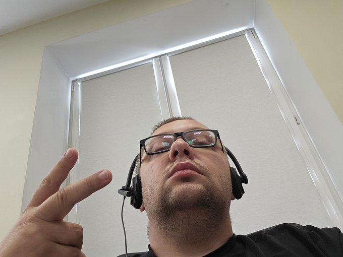 A low-angle selfie of a young man wearing black-rimmed glasses and over-ear headphones, making a peace sign with his hand.