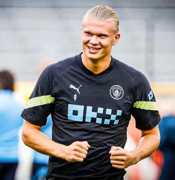 Erling Haaland smiling while wearing a black Puma training jersey with 'OKX' sponsor and Manchester City crest during what appears to be a training session.