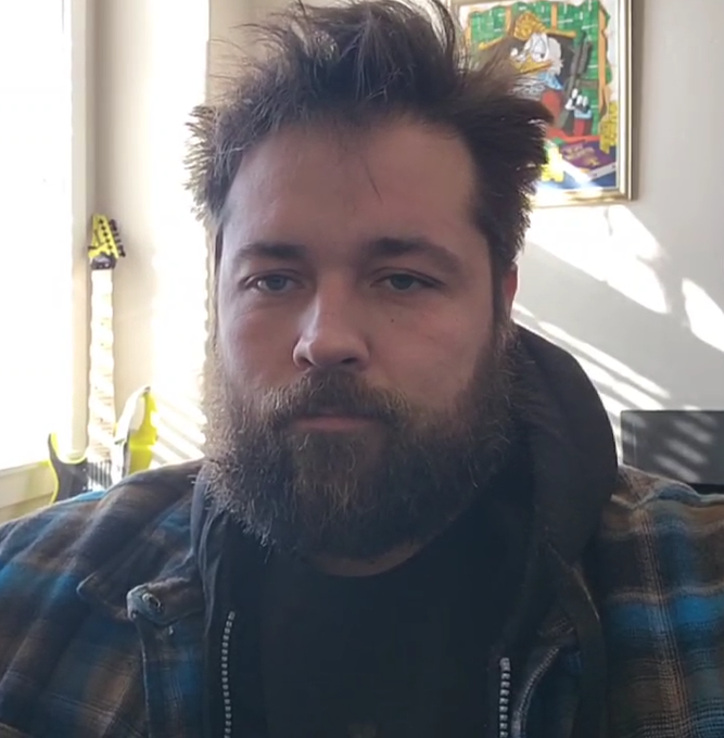 A selfie of a man with a beard and messy hair wearing a plaid shirt, with a guitar and artwork visible in the background.
