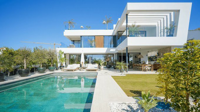 A modern white luxury villa featuring a swimming pool, outdoor lounge area, and landscaped gardens under a clear blue sky.