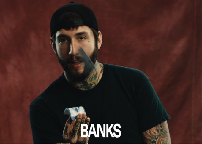 A heavily tattooed man wearing a black cap and t-shirt holds a knife in a menacing pose against a deep red backdrop, with the name "BANKS" displayed in white text at the bottom of the frame. The image appears to be a still from a music video or visual for an artist named Banks, paired with the post's caption referencing crypto market gains ("hpos up 50%") and the grind of trying to find alpha ("lock in onchain")—the aggressive, cutthroat aesthetic of the imagery mirroring the competitive, predatory tone of chasing profits in crypto.