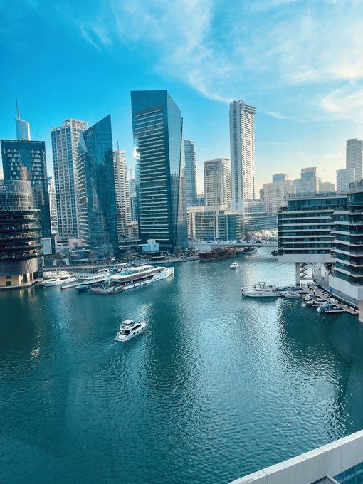A daytime view of Dubai Marina shows turquoise waters with boats and yachts, surrounded by modern high-rise buildings under a blue sky.