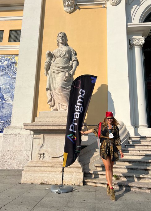 A woman wearing a red cap, sunglasses, and a patterned dress stands on a step next to a statue, holding a black flag that reads 'Pragma' with a colorful logo.