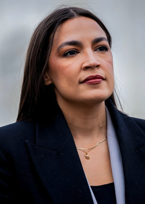 Portrait of a woman with long dark hair wearing a black blazer and a gold necklace with a teardrop pendant, looking off to the side.