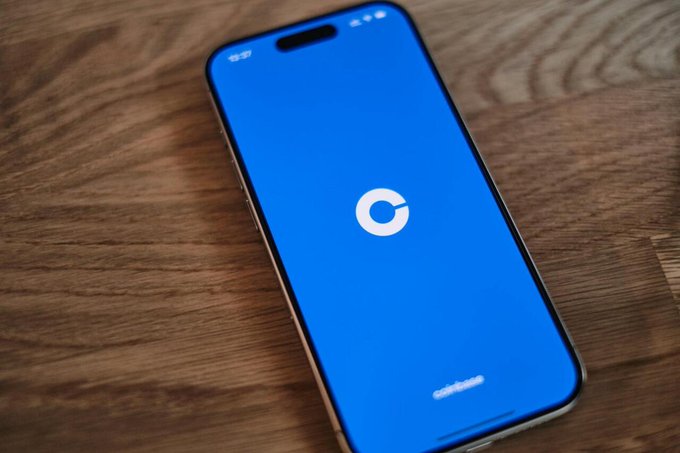 An iPhone lies on a wooden surface displaying the blue Coinbase app logo with "coinbase" text at the bottom of the screen.