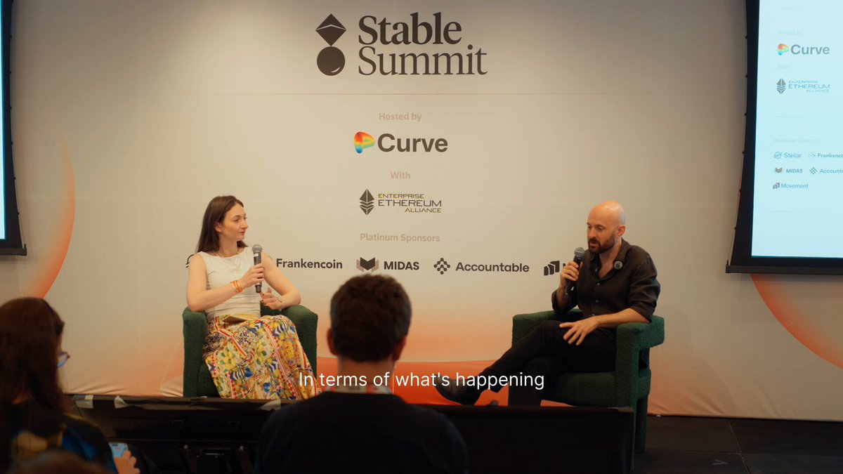 The image shows a man and a woman sitting next to each other, engaged in conversation. They are positioned behind a podium that displays text related to the "Stable Summit" event. The background features an orange wall with various logos and text, including one for "Curve," which is likely associated with the event or company they are discussing.