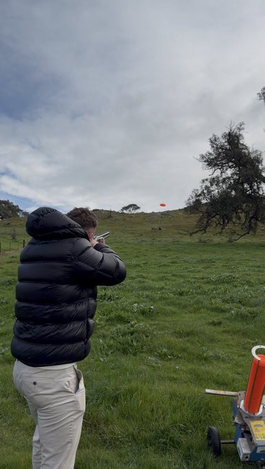A person in a black puffer jacket and light pants aiming what appears to be a launcher in a grassy field under a cloudy sky.