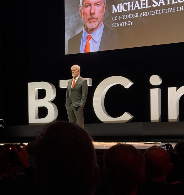 A man in a dark suit and orange tie stands on stage at a Bitcoin conference, with his photo and the title 'MICHAEL SAYLOR, CO-FOUNDER AND EXECUTIVE CHAIRMAN, STRATEGY' displayed on a large screen behind him.