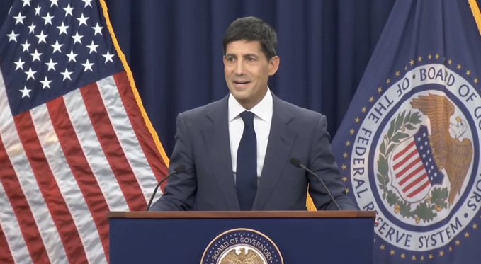 A man in a navy suit speaks at a podium bearing the Board of Governors seal, with the American flag and Federal Reserve System flag displayed behind him.