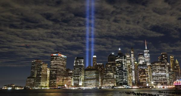 A nighttime cityscape of the New York City skyline featuring the Tribute in Light memorial beams shining into the cloudy sky above the illuminated skyscrapers.