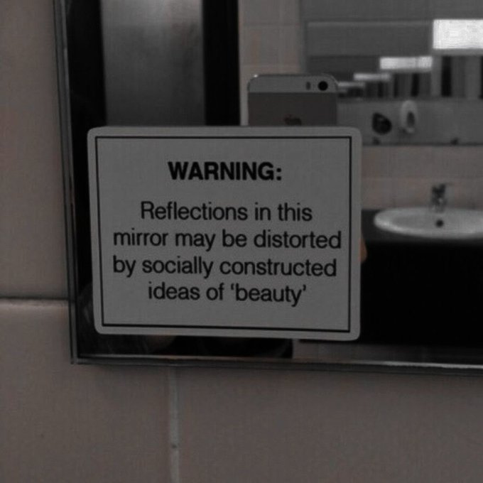 A sign affixed to a bathroom mirror reads: "WARNING: Reflections in this mirror may be distorted by socially constructed ideas of 'beauty'."
