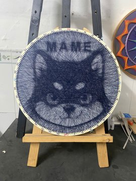 A piece of string art depicting a black cat's face with the name "MAME" on an embroidery hoop displayed on a small easel.