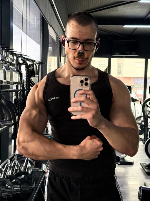 A muscular man with glasses and a nose-piercing taking a mirror selfie in a gym, wearing a black tank top and flexing his arm.