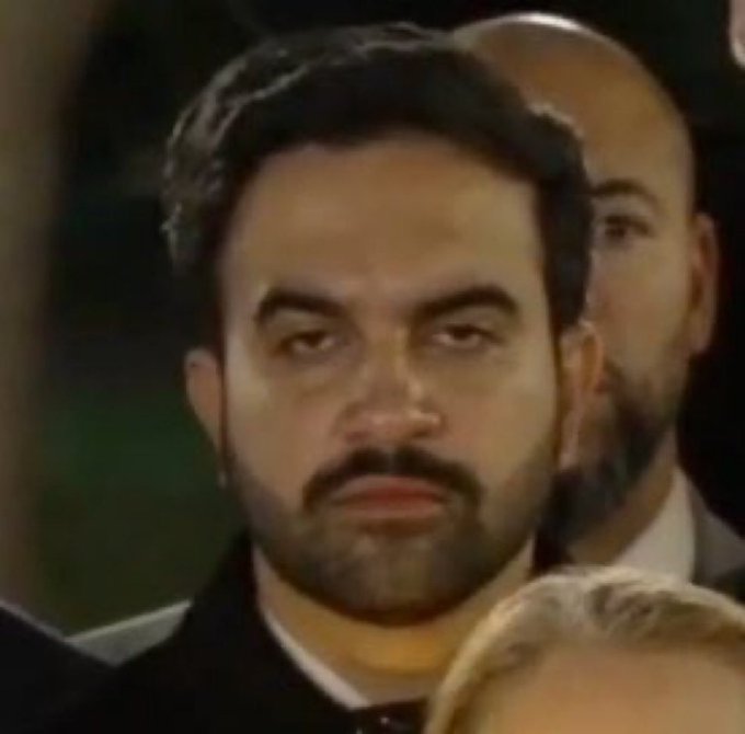 A close-up shot of a man with dark hair and a beard wearing a dark suit, standing in what appears to be a crowded event.