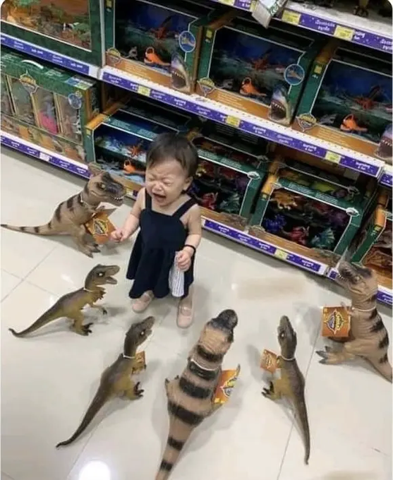 A crying toddler in a black dress stands on a tiled floor holding a dinosaur toy, surrounded by several other lifelike dinosaur toys that appear to be looking at her.