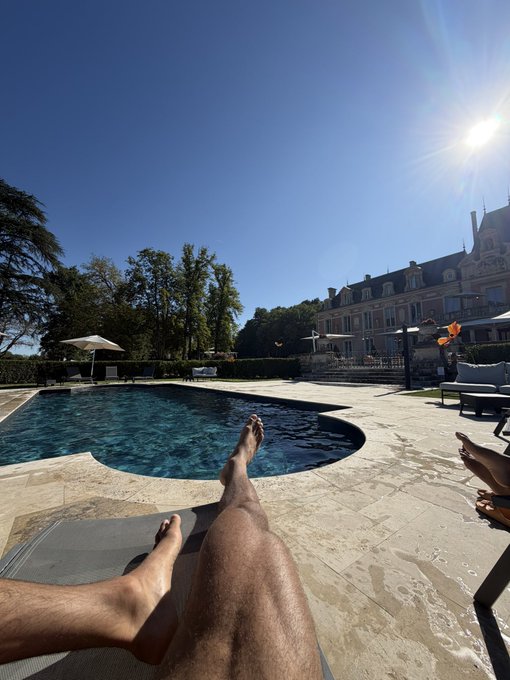 First-person perspective of someone relaxing on a lounge chair by a swimming pool, with their legs extended toward the water, set against a grand estate building under a bright sunny sky.