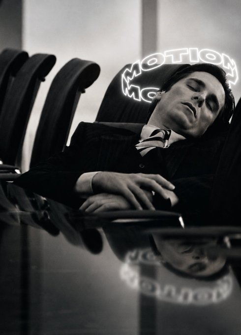 A black and white surreal photo of a man in a suit sleeping with his head resting on chairs, with a neon sign reading 'MOTION' reflected in the glossy surface below.