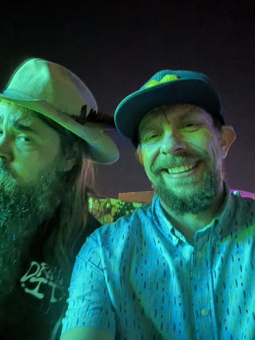 Two men wearing hats, one in a wide-brimmed tan hat and the other in a baseball cap with a teal shirt, smile under colorful green and blue stage lighting.