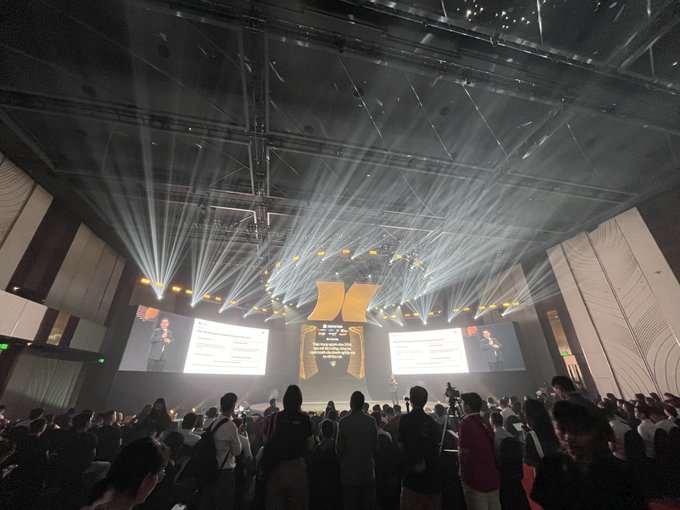 A photograph of a large conference event with dramatic stage lighting, multiple projection screens, and an audience watching a presentation in a modern auditorium.