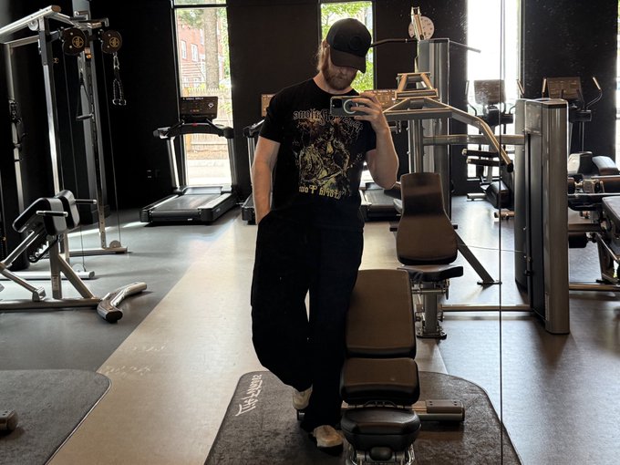A man in a black cap and graphic t-shirt takes a mirror selfie in a gym, wearing black pants, surrounded by workout equipment and treadmills.
